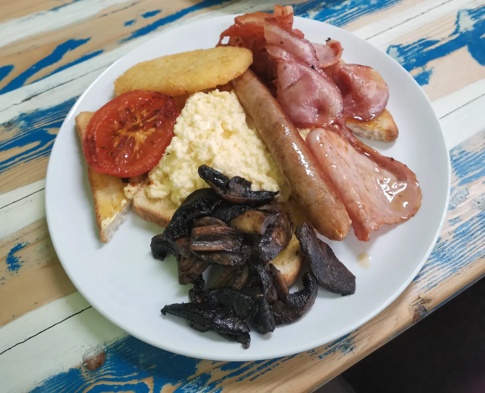 Full cooked breakfast plate with eggs, bacon, sausage and tomato on painted timber table
