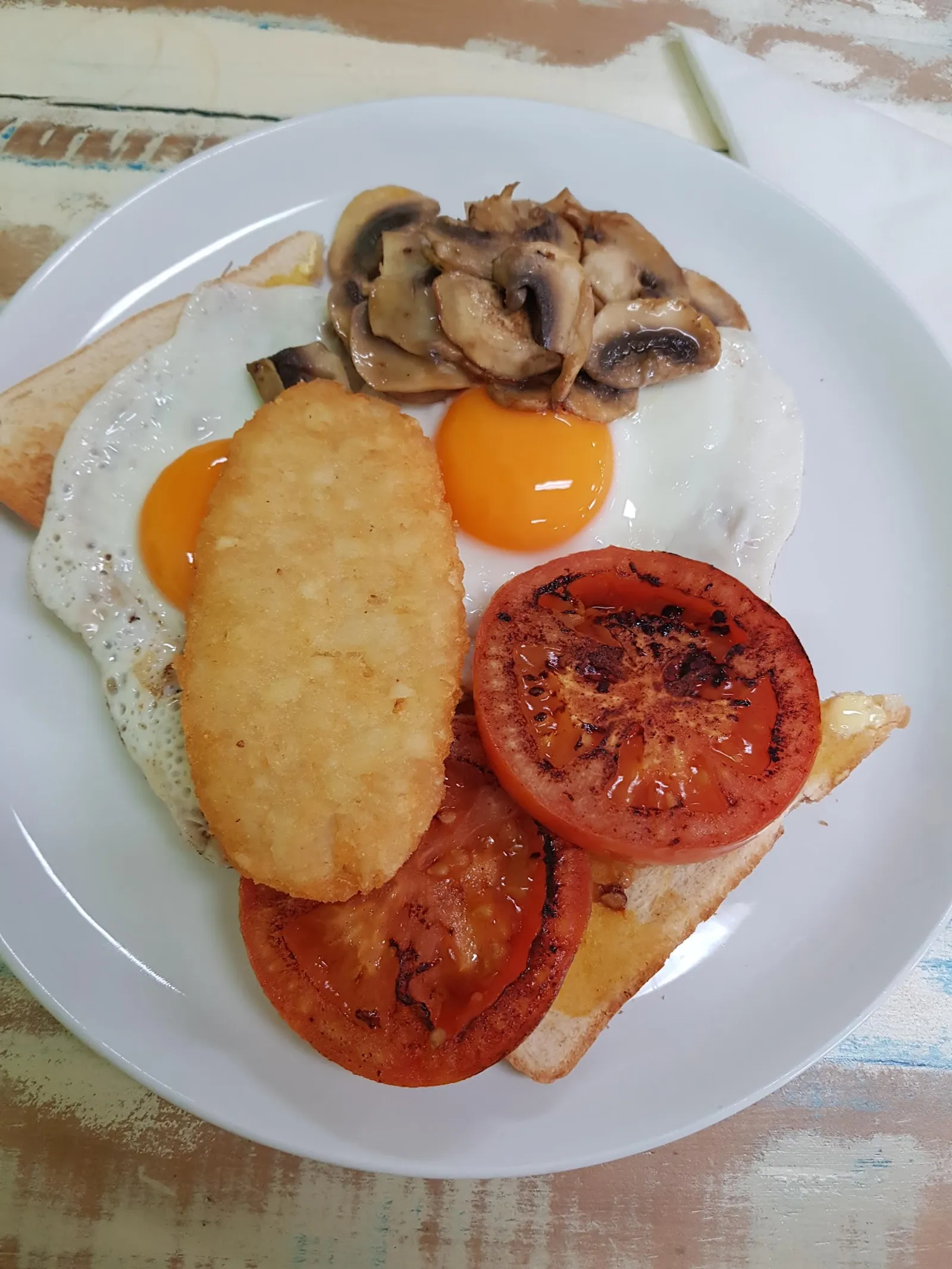 Breakfast plate with fried egg, hash brown, tomato and mushrooms on painted timber table
